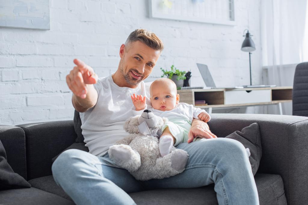 padre sonriente sentado en el sofá con la hija bebé y apuntando con el dedo a algo
 - Foto, imagen