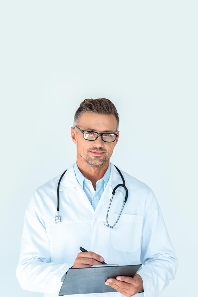 pohledný doktor v brýlích s stetoskop na bedrech napsat něco do schránky a při pohledu na fotoaparát izolované na bílém - Fotografie, Obrázek