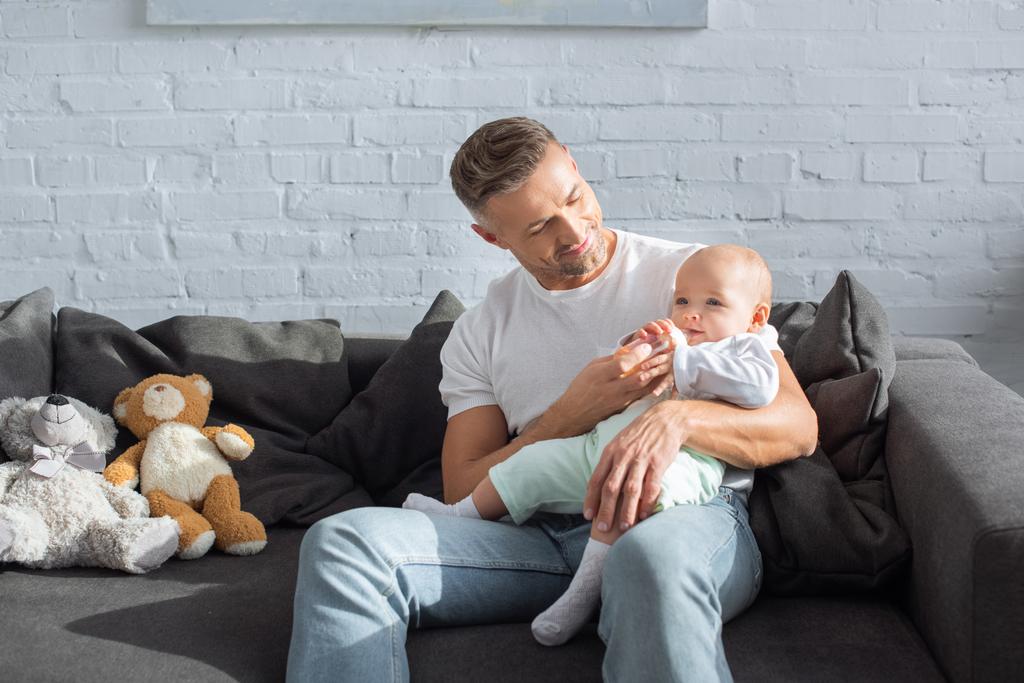 smiling father sitting on couch and feeding baby daughter in living room - Photo, Image