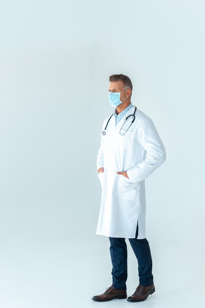 médico adulto con bata blanca y máscara médica mirando hacia otro lado aislado en blanco
 - Foto, imagen