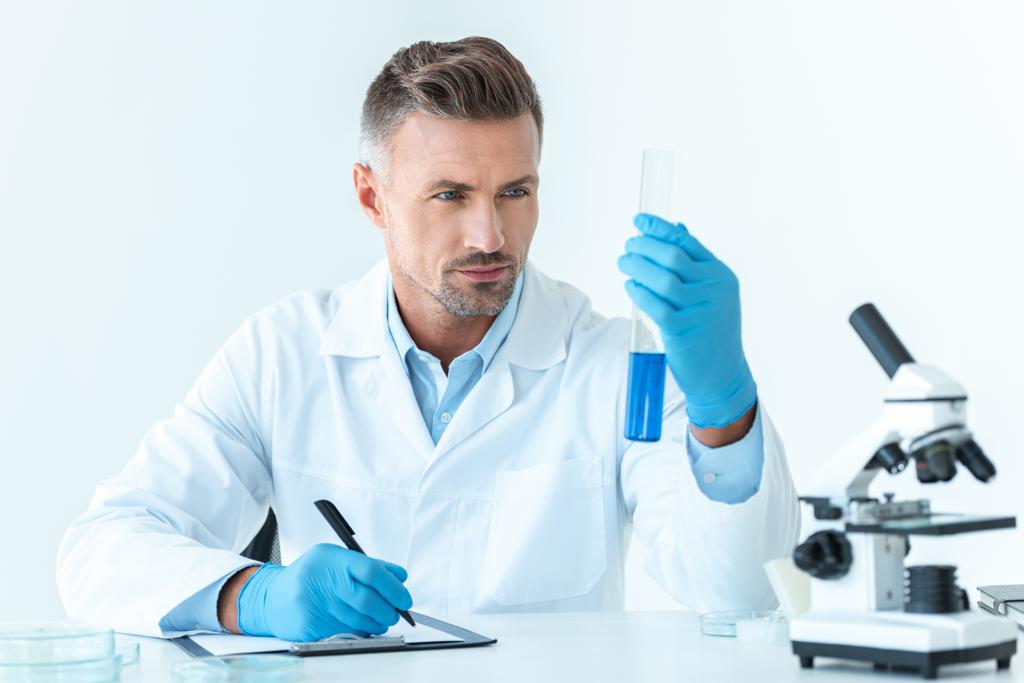 красивый ученый смотрит на пробирку с голубым реагентом, изолированным на белом
 - Фото, изображение