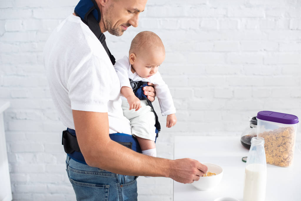 father holding infant daughter in baby carrier and preparing breakfast  - Photo, Image