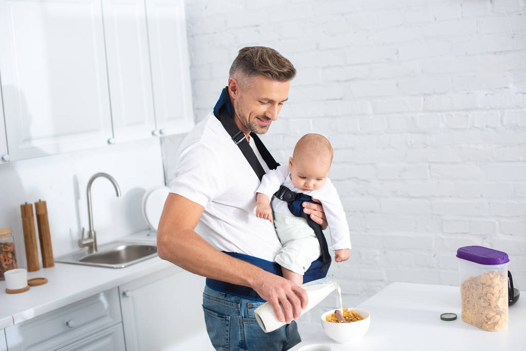 Vater hält kleine Tochter in Tragetasche und gießt Milch in Schüssel mit Cornflakes  - Foto, Bild