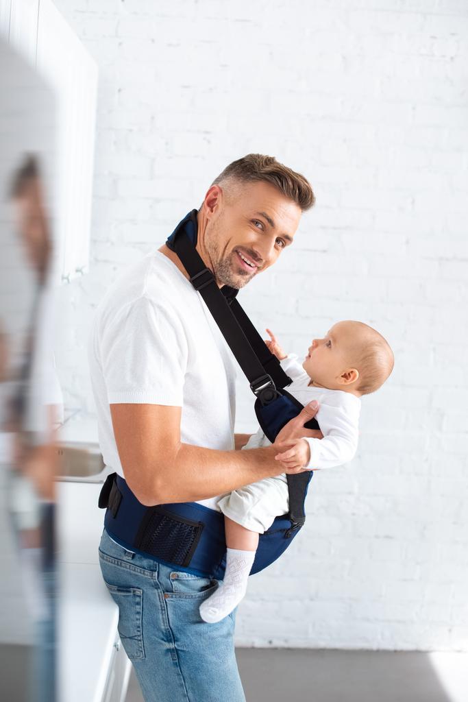 cheerful father holding infant daughter in baby carrier at home  - Photo, Image