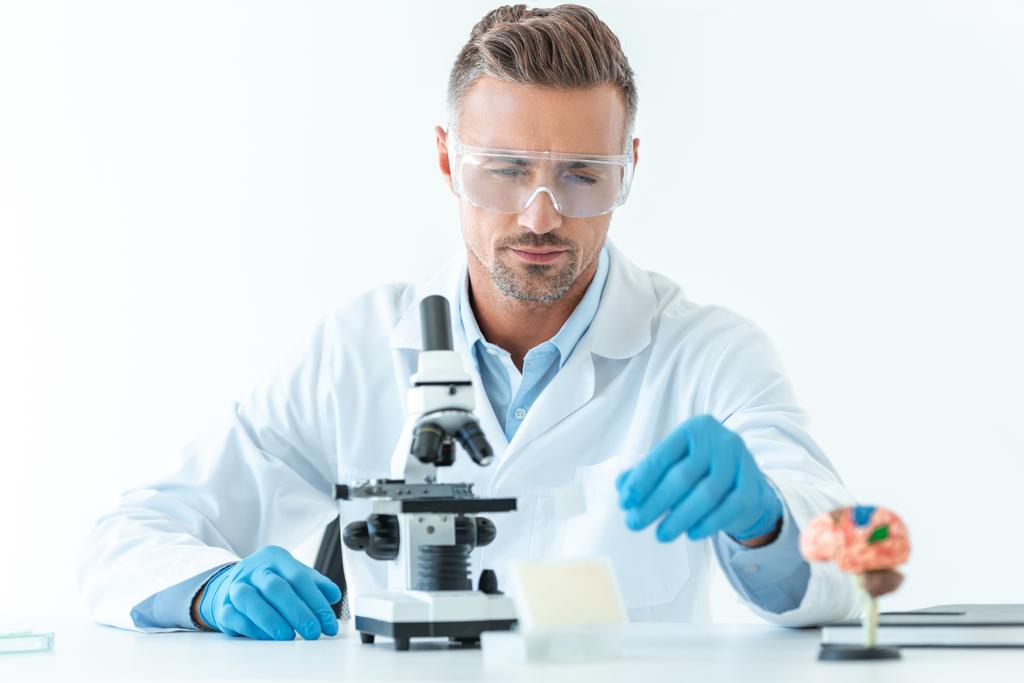 Selektywny fokus przystojny naukowca w ochronne okulary podejmowania eksperyment z mikroskopu na białym tle - Zdjęcie, obraz