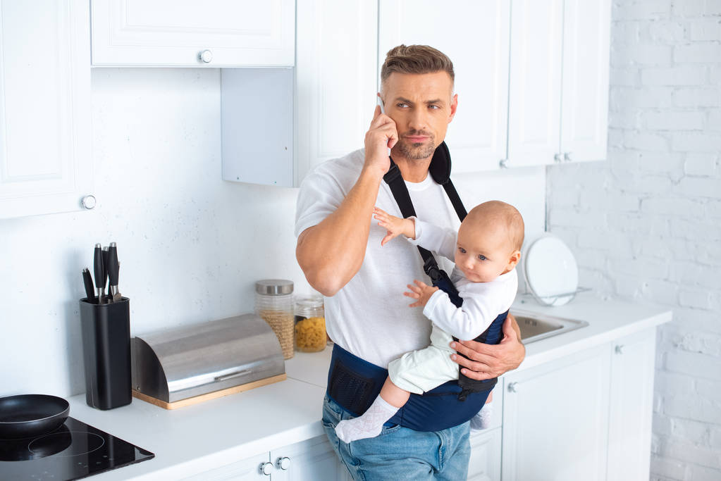 otec drží dcerkou v nosítko a mluví na smartphone v kuchyni - Fotografie, Obrázek