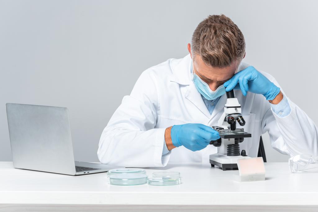 schöner Wissenschaftler mit medizinischer Maske und medizinischen Handschuhen, der das Mikroskop isoliert auf weißem Grund betrachtet - Foto, Bild