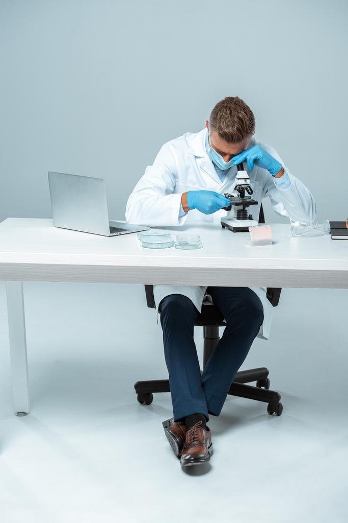schöner Chemiker mit medizinischer Maske und medizinischen Handschuhen, der das Mikroskop isoliert auf weißem Grund betrachtet - Foto, Bild