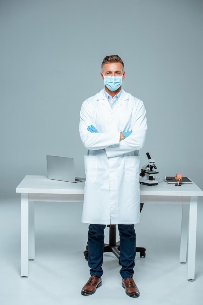 guapo científico en máscara médica y guantes médicos de pie con los brazos cruzados aislados en blanco
 - Foto, Imagen