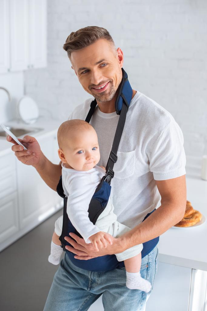 iloinen isä, joka käyttää älypuhelinta ja pitää lapsen tytärtä vauvan kantoaallossa
  - Valokuva, kuva