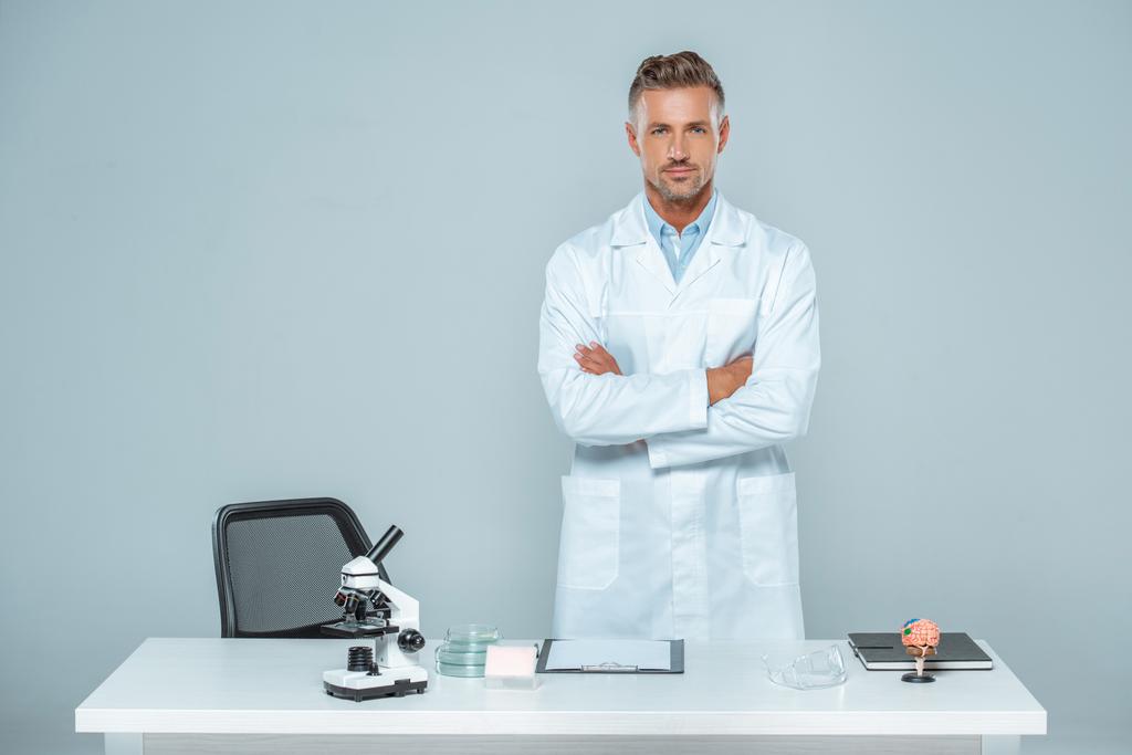 όμορφος επιστήμονας στέκεται με σταυρωμένα τα χέρια κοντά στο τραπέζι που απομονώνονται σε λευκό - Φωτογραφία, εικόνα