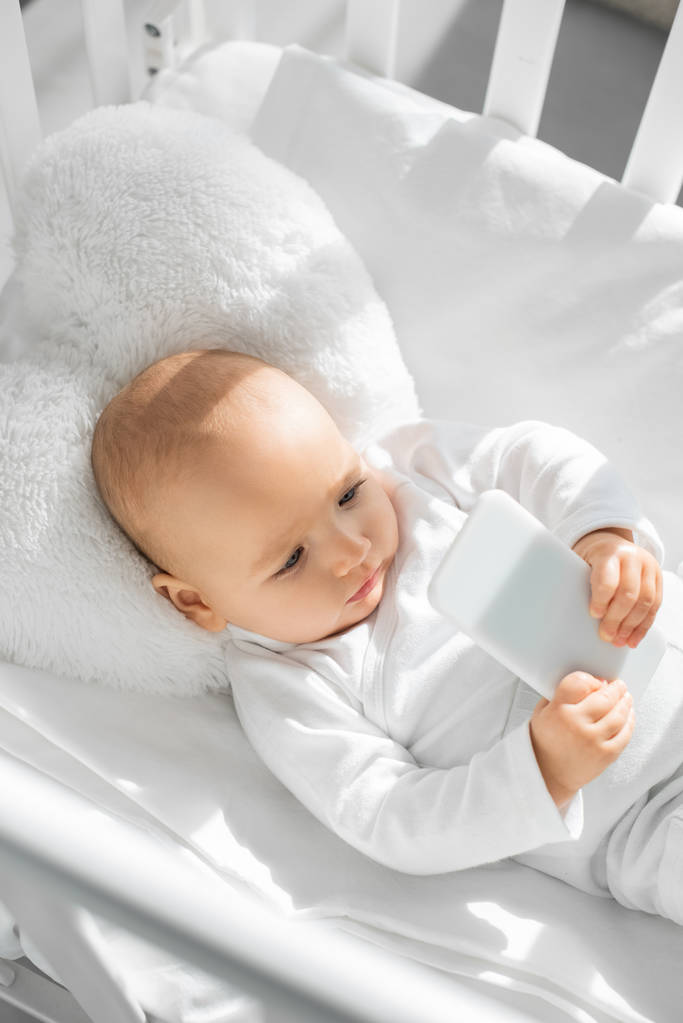 cute infant kid holding smartphone in white baby crib  - Photo, Image