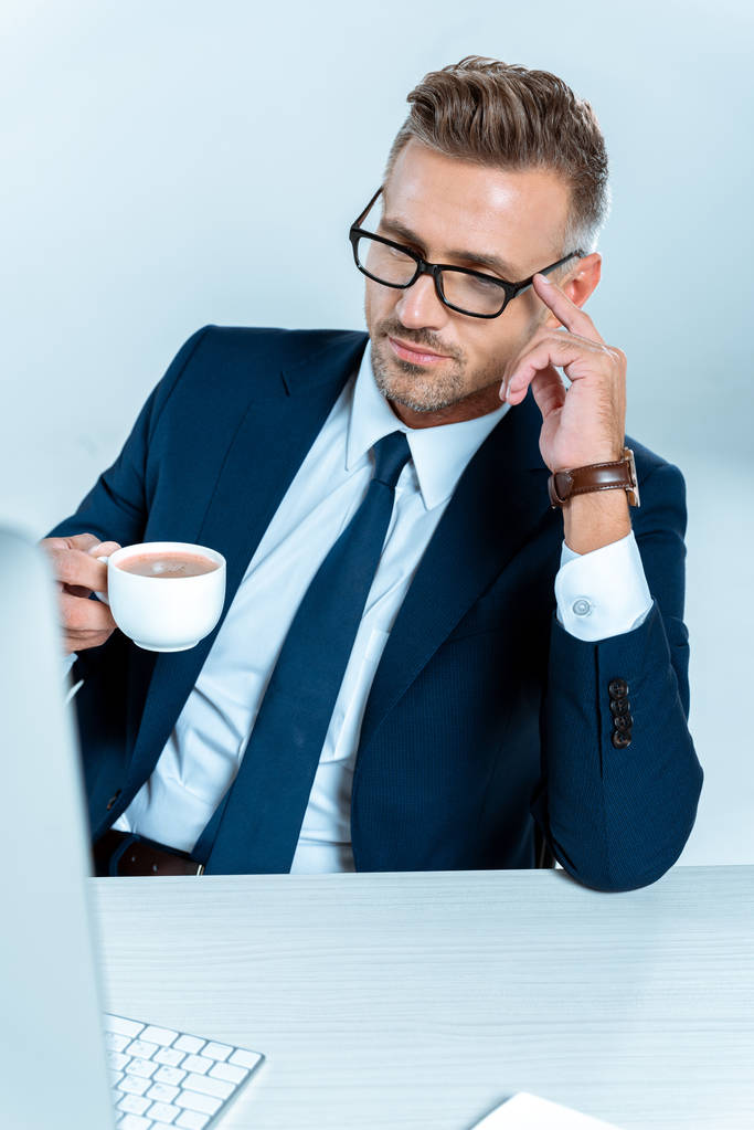 guapo hombre de negocios sosteniendo la taza de café y mirando a la computadora aislada en blanco
 - Foto, imagen