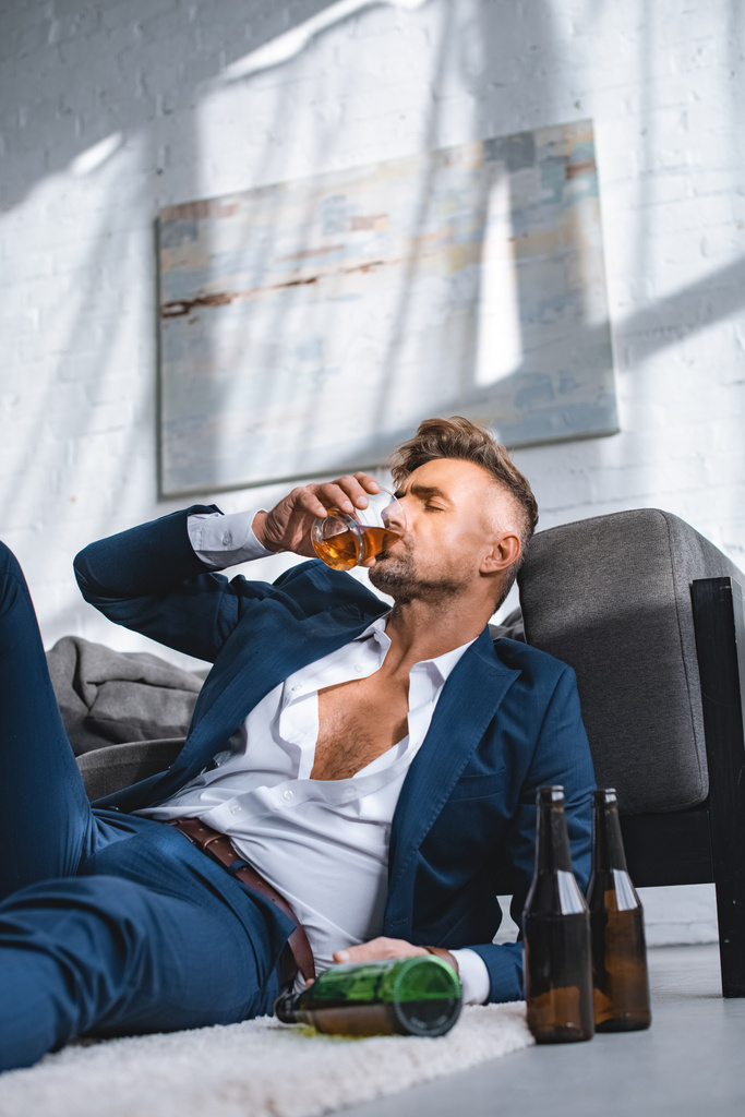 Depressiver Geschäftsmann trinkt Whiskey in der Nähe von Flaschen im Wohnzimmer  - Foto, Bild
