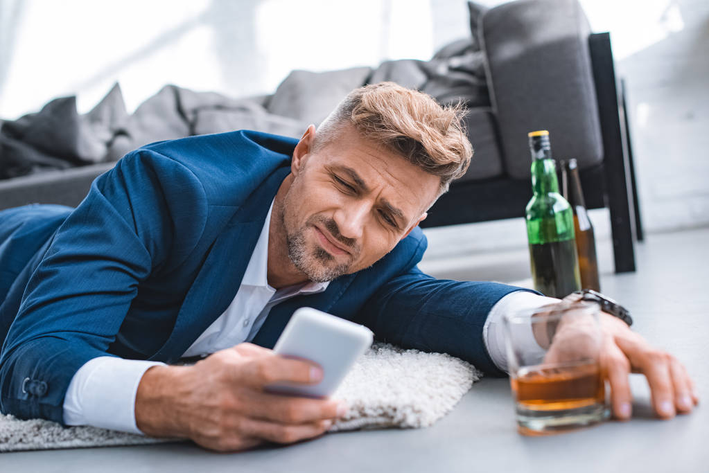 enfoque selectivo de hombre de negocios borracho acostado en la alfombra y el uso de teléfonos inteligentes
   - Foto, Imagen