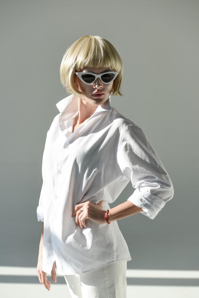 attraktive blonde Frau mit Sonnenbrille und modischem weißen Outfit mit Handakimbo auf weißem Hintergrund - Foto, Bild