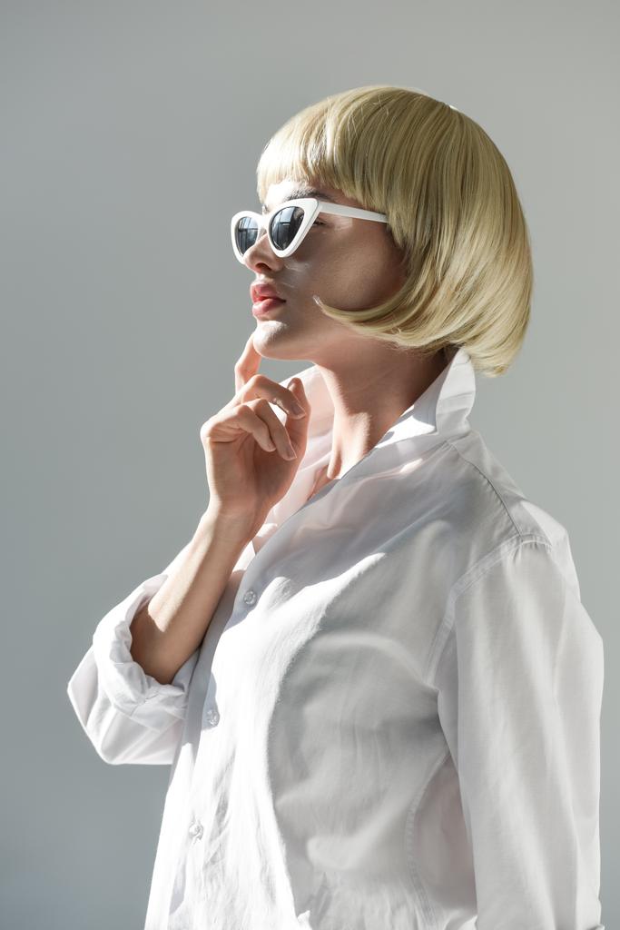 Zijaanzicht van aantrekkelijke blonde vrouw in zonnebrillen en modieuze witte outfit aan kin op wit wordt geïsoleerd te raken - Foto, afbeelding