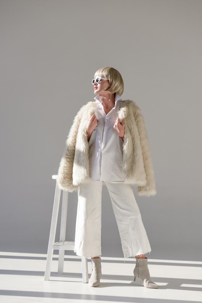 привлекательная блондинка в солнцезащитных очках и модном зимнем наряде с искусственной шубой, смотрящая на белый
 - Фото, изображение