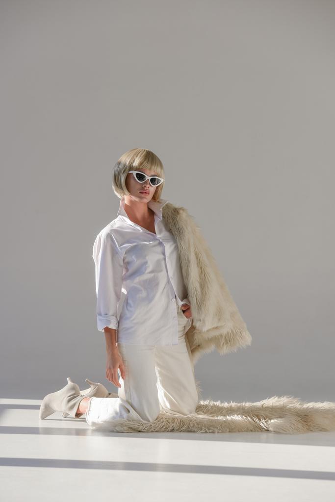 mujer atractiva en gafas de sol y ropa de invierno de moda con abrigo de piel sintética de pie sobre las rodillas en la alfombra en blanco
 - Foto, imagen