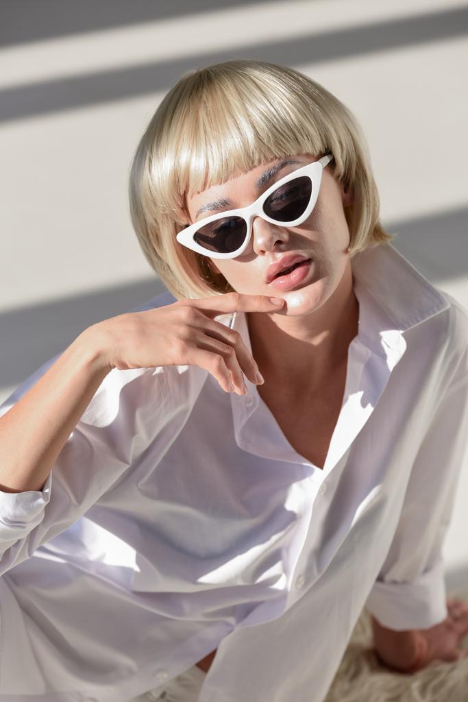 vue grand angle de femme blonde sensuelle dans des lunettes de soleil et tenue d'hiver à la mode assis sur manteau en fausse fourrure sur blanc
 - Photo, image