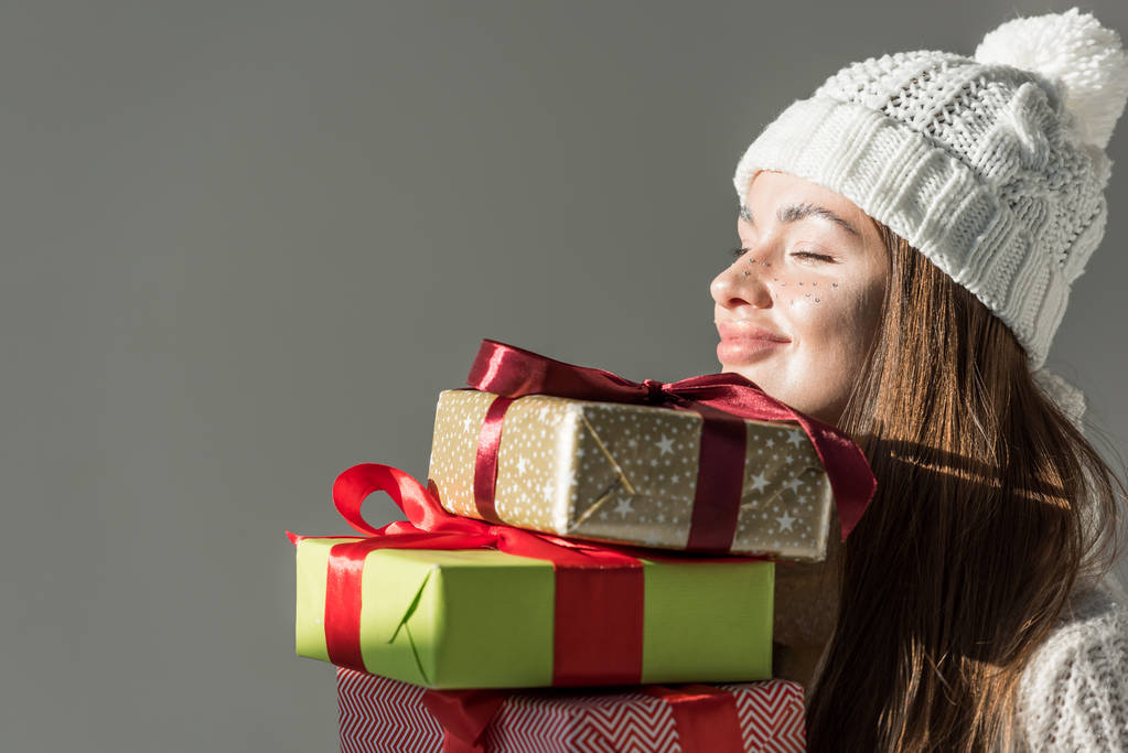 cheerful attractive woman in stylish winter sweater and scarf holding gift boxes isolated on grey - Photo, Image