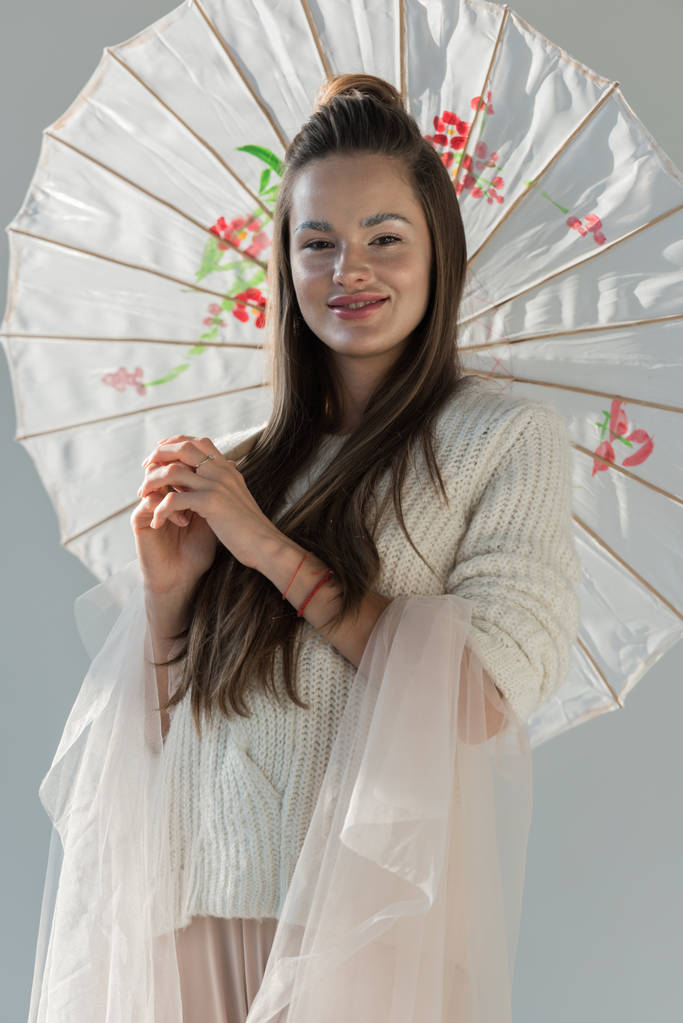 portrait of attractive woman in fashionable winter outfit standing with paper umbrella and looking at camera on white - Photo, Image