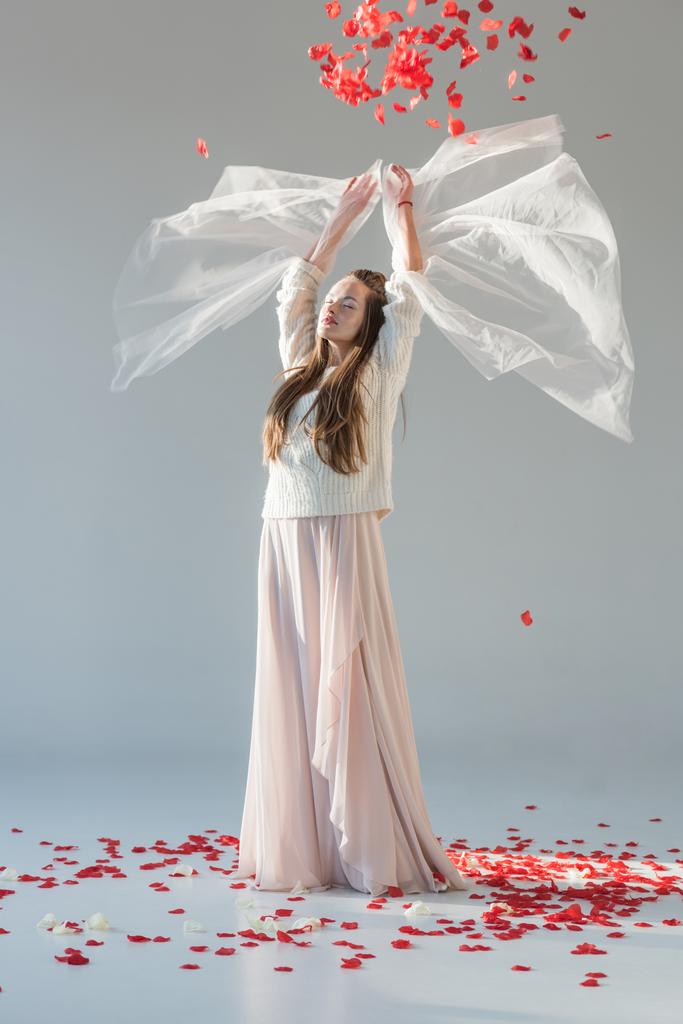 mulher atraente em roupa de inverno na moda em pé com as mãos levantadas sob queda pétalas de rosas vermelhas isoladas em branco
 - Foto, Imagem