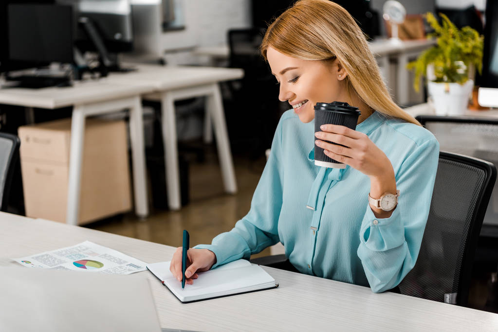 uśmiecha się kobieta z kawą do przejść robiąc notatki w notesie w miejscu pracy w biurze - Zdjęcie, obraz