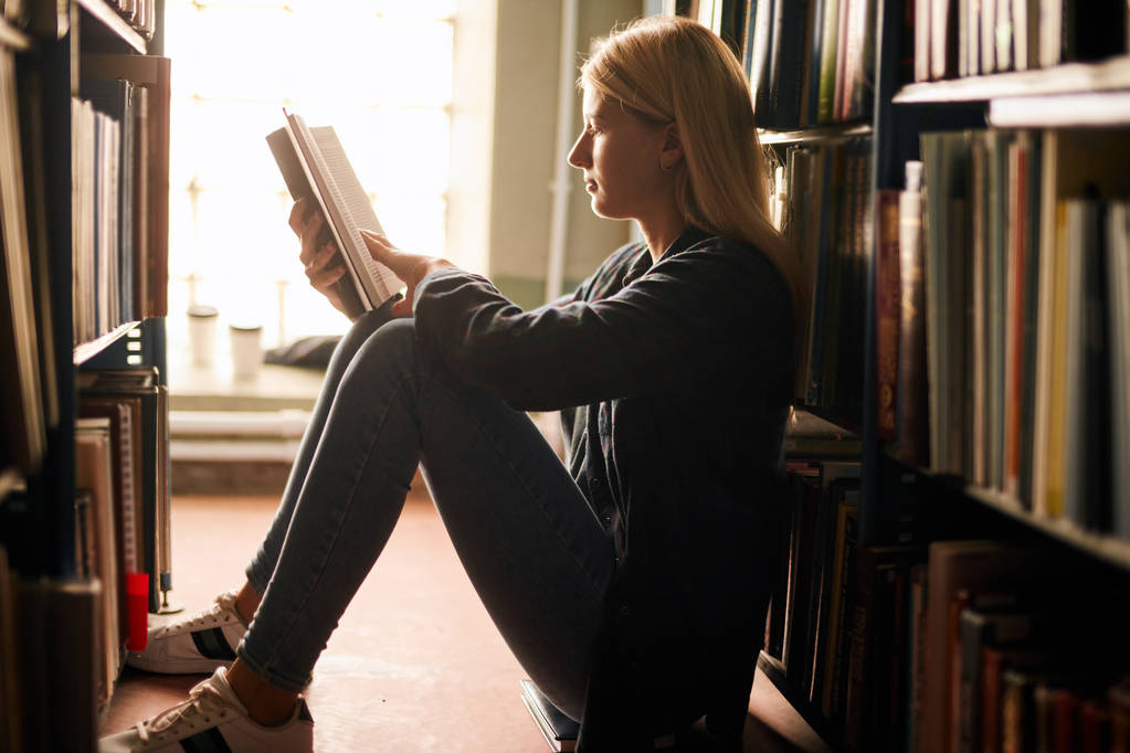 Νεαρό κορίτσι που κάθεται στο πάτωμα στη βιβλιοθήκη μεταξύ ράφια, διαβάζοντας βιβλία. - Φωτογραφία, εικόνα