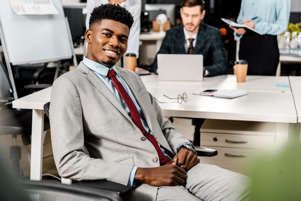вибірковий фокус усміхненого афроамериканського бізнесмена та колег позаду на робочому місці в офісі
 - Фото, зображення