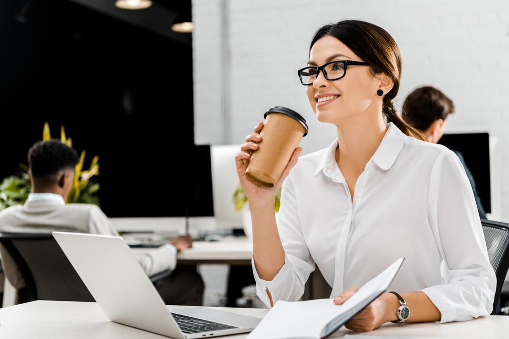 joven empresaria en gafas con café para llevar y cuaderno en el lugar de trabajo con portátil en la oficina
 - Foto, Imagen
