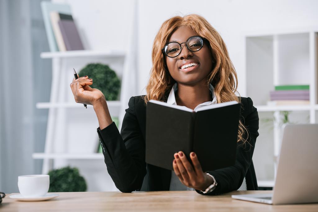 mujer de negocios afroamericana feliz sosteniendo pluma y cuaderno
  - Foto, Imagen