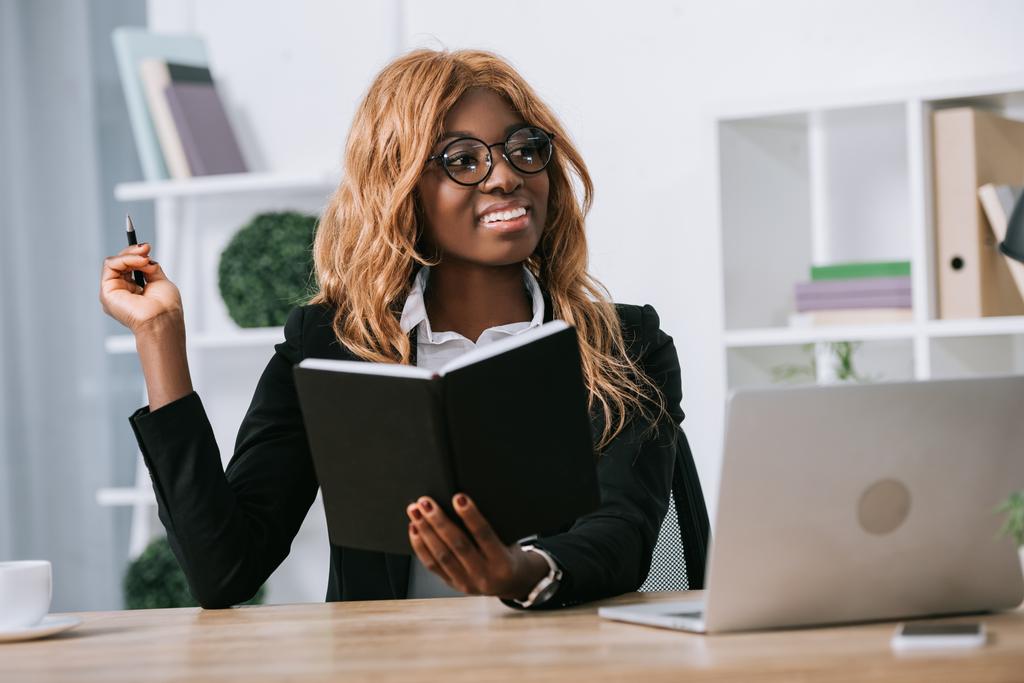 счастливая африканская американская бизнесвумен сидит в очках и держит ноутбук и ручку
  - Фото, изображение