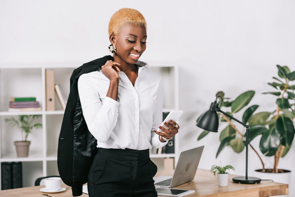 afrikanisch-amerikanische Geschäftsfrau mit kurzen Haaren blickt auf Smartphone und hält Jacke  - Foto, Bild