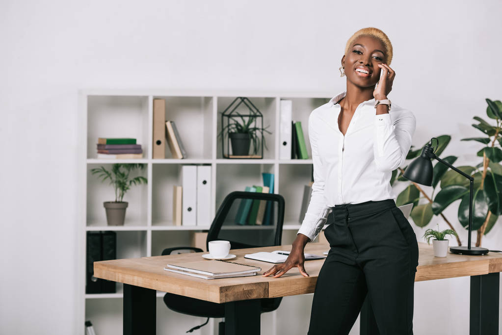 glückliche afrikanisch-amerikanische Geschäftsfrau mit kurzen Haaren spricht im modernen Büro auf dem Smartphone - Foto, Bild