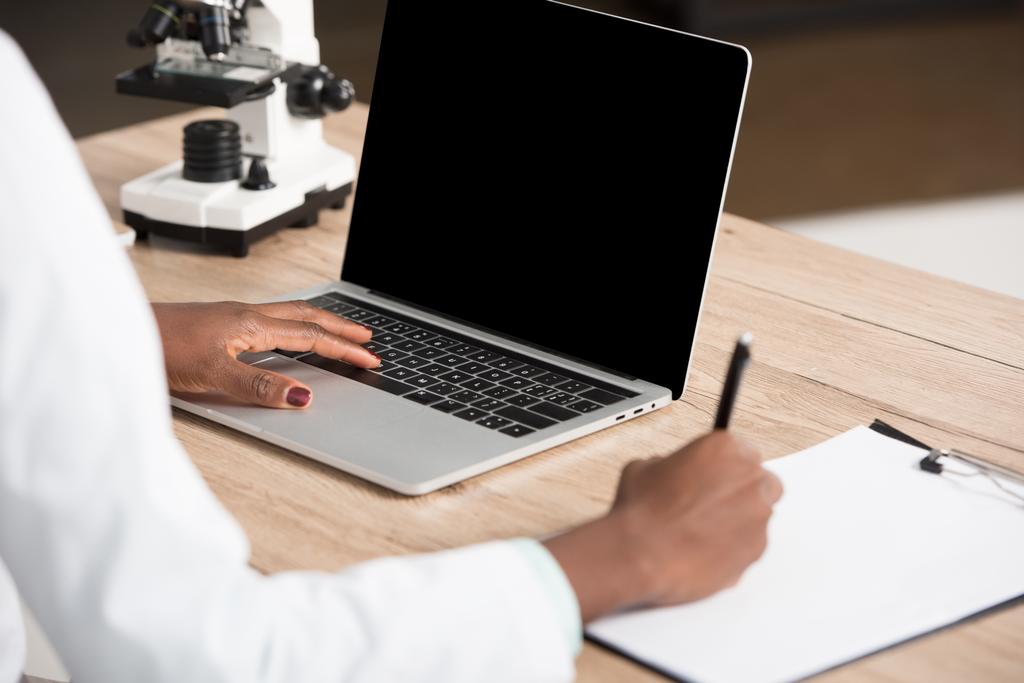 vue recadrée du médecin afro-américain écrivant sur papier et utilisant un ordinateur portable avec écran blanc
  - Photo, image