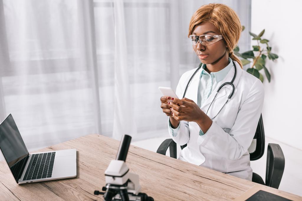 aantrekkelijke Afro-Amerikaanse wetenschapper zitten in bril en het gebruik van smartphone in laboratorium - Foto, afbeelding