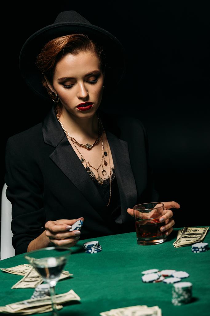 atractiva chica en chaqueta y sombrero sosteniendo vaso de whisky y fichas de póquer en la mesa en el casino
 - Foto, Imagen