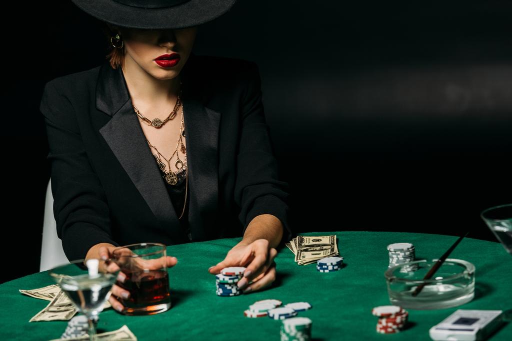 ελκυστική τζογαδόρος με σακάκι και καπέλο κρατά ποτήρι ουίσκι στο τραπέζι του πόκερ στο καζίνο - Φωτογραφία, εικόνα