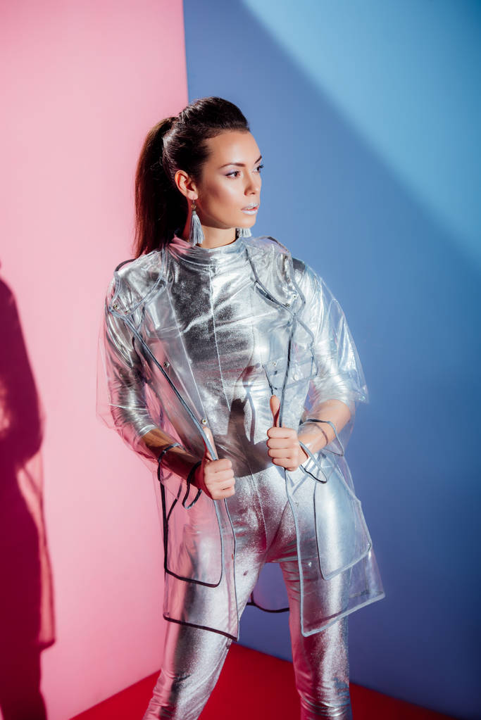beautiful stylish woman posing in silver bodysuit and raincoat on pink and blue background - Photo, Image