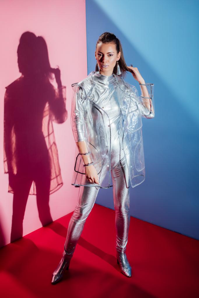 vonzó elegáns nő pózol ezüst Body és esőkabátot, rózsaszín és kék háttér - Fotó, kép
