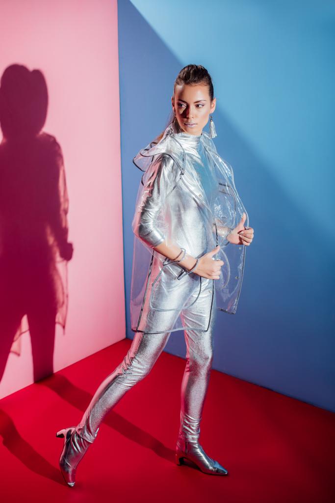 elegant stylish young woman posing in silver bodysuit and raincoat on pink and blue background - Photo, Image