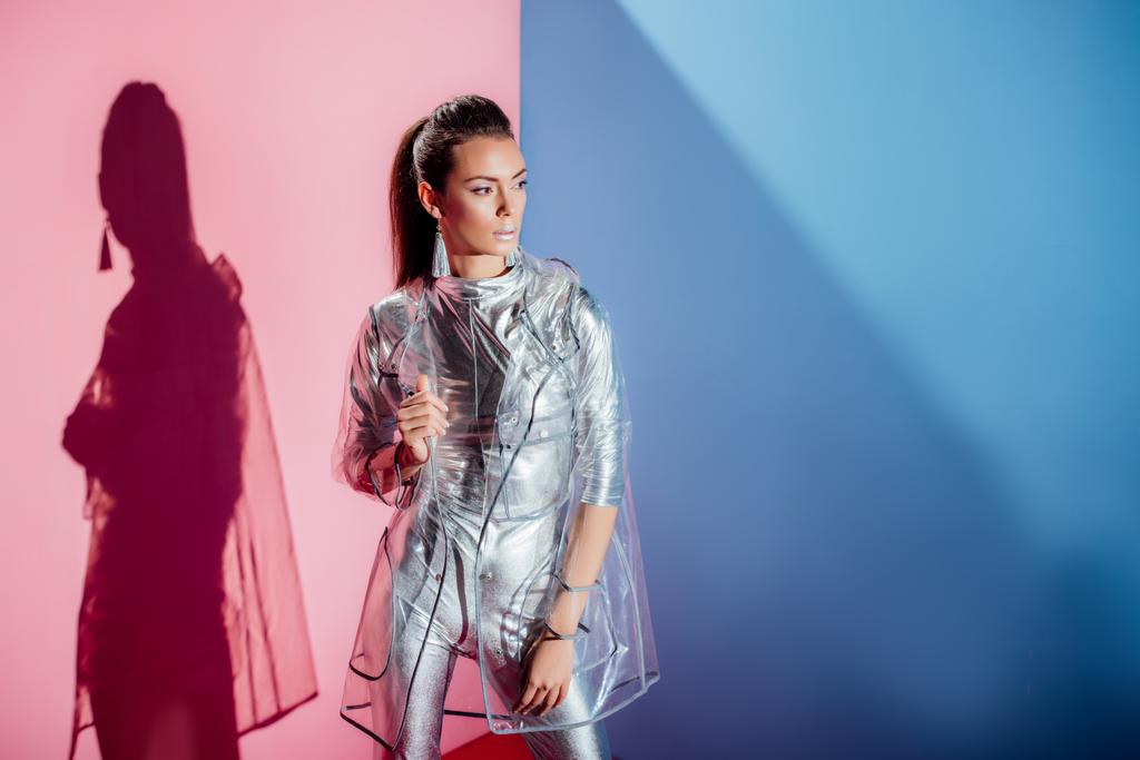 beautiful girl posing in metallic bodysuit and trendy raincoat on pink and blue background - Photo, Image