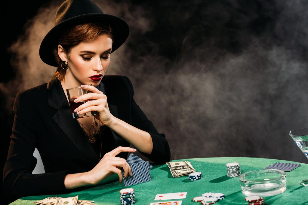 nadenkend aantrekkelijk meisje in jas en hoed pokeren en houden van glas whiskey in casino - Foto, afbeelding