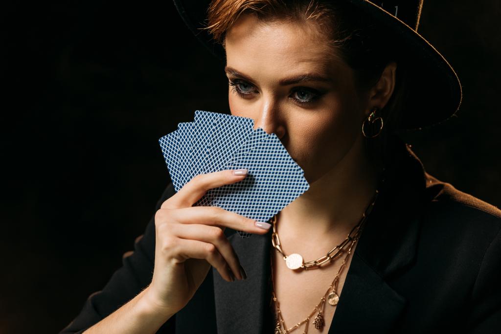 attraktives Mädchen in Jacke und Hut, das Gesicht mit Pokerkarten verdeckt, isoliert auf schwarz und wegschauend - Foto, Bild