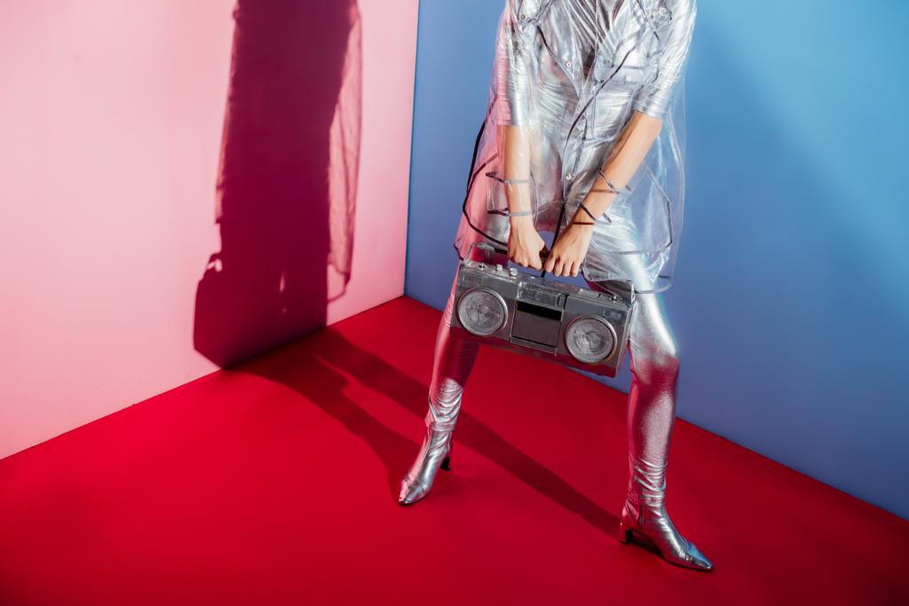обрізаний вид жінки в металевому костюмі для тіла та плащі, позує з бумбокс на рожевому та синьому фоні
 - Фото, зображення