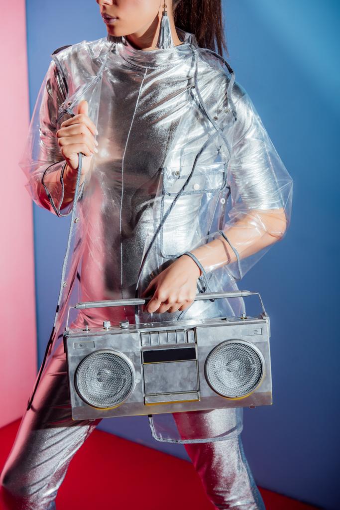 cropped view of girl in silver bodysuit and raincoat posing with boombox on pink and blue background - Photo, Image