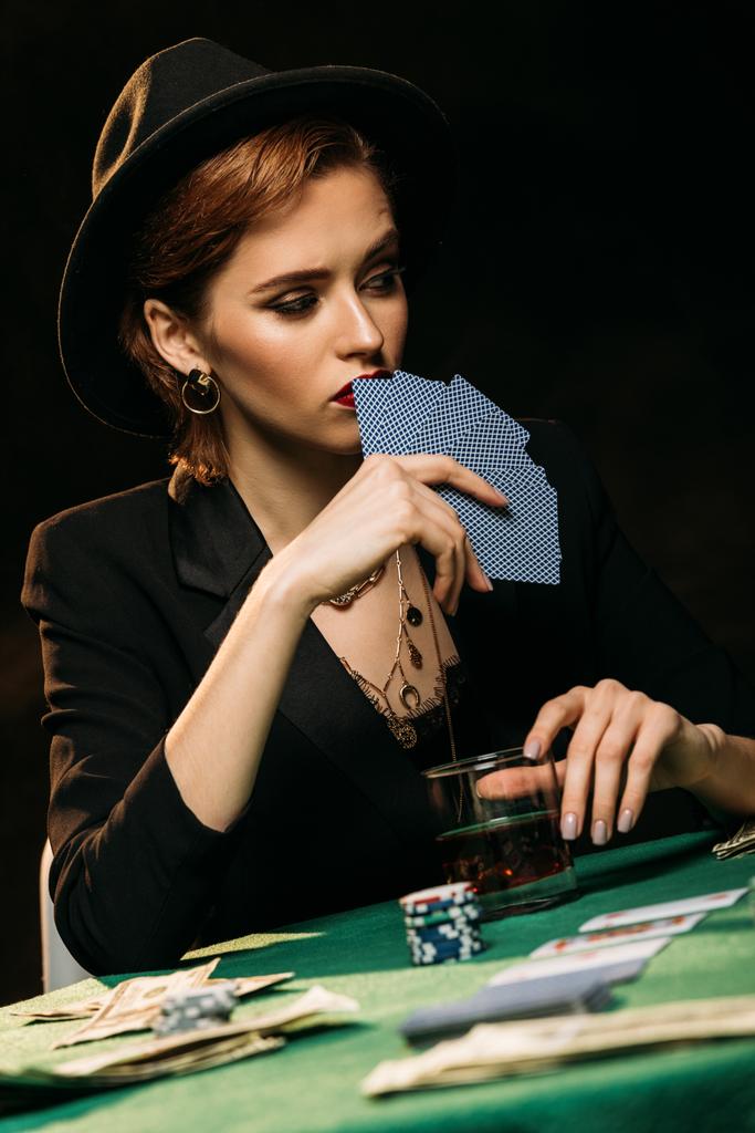 surface level of attractive girl in jacket and hat covering face with poker cards in casino - Photo, Image