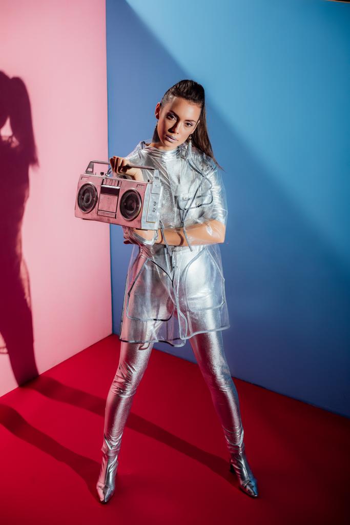 beautiful young woman in metallic bodysuit and raincoat posing with boombox on pink and blue background - Photo, Image