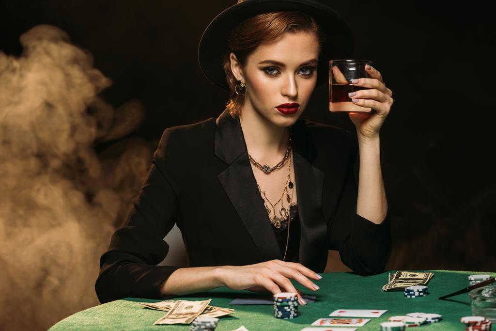 attraktives Mädchen in Jacke und Hut hält ein Glas Whiskey am Pokertisch im Casino und blickt in die Kamera - Foto, Bild